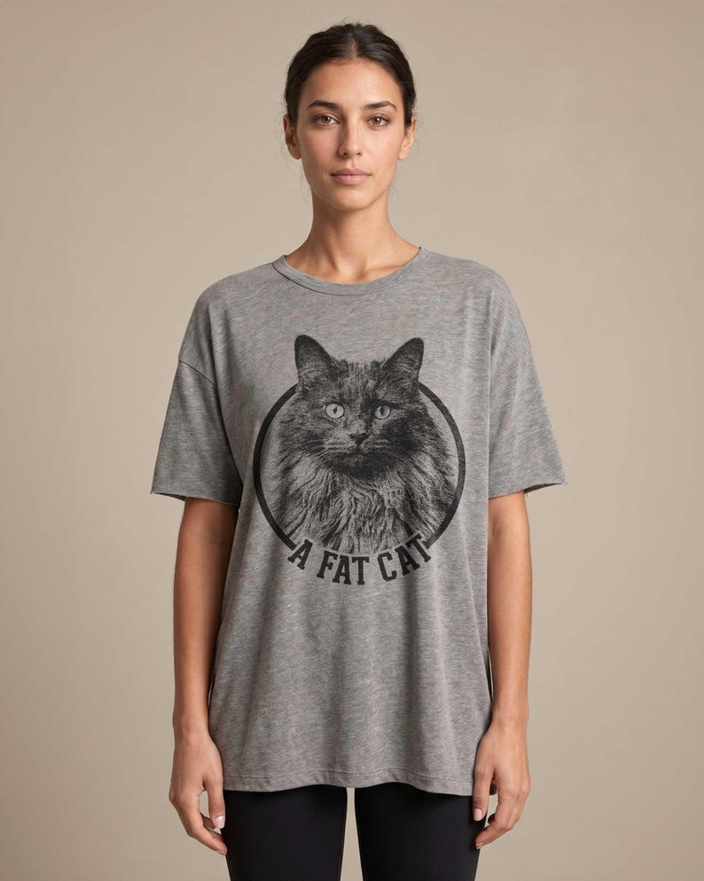Puede incluir: Camiseta gris con una ilustraci&oacute;n en blanco y negro de un gato peludo dentro de un c&iacute;rculo negro. El texto "A FAT CAT" est&aacute; impreso debajo de la imagen del gato. La camiseta tiene mangas cortas y un corte holgado.