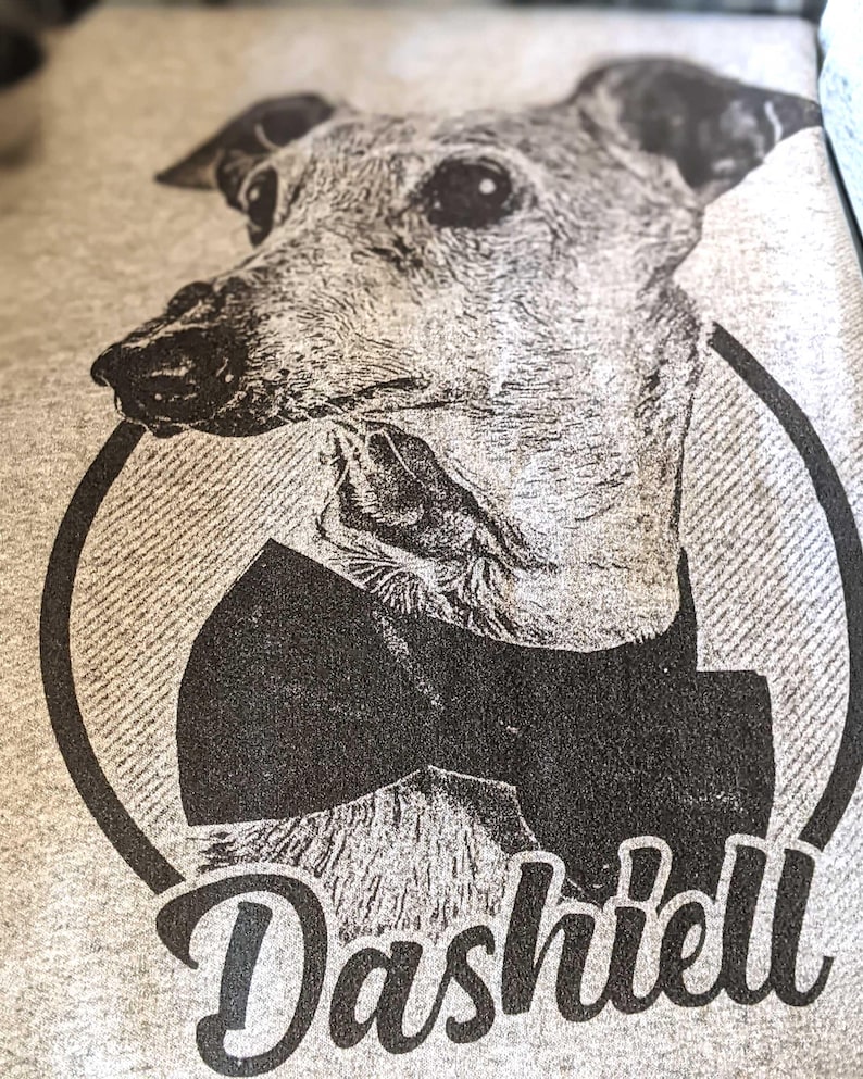 May include: A heather gray t-shirt featuring a black ink graphic of a greyhound dog wearing a bow tie inside a circular frame. The name Dashiell is printed in a bold, cursive script font beneath the circular graphic of the dog.