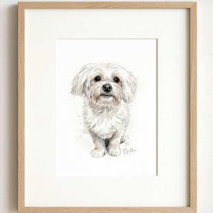 May include: A watercolor painting of a small white dog with floppy ears and dark eyes, centered on a white background. The artwork is enclosed in a light-colored wooden frame with a white mat. The name Millie is written in cursive at the bottom.
