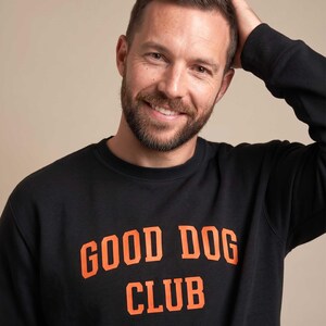 May include: A black long-sleeve sweatshirt featuring the text GOOD DOG CLUB printed in bold orange capital letters across the chest. The garment has a classic crew neckline and long sleeves, constructed from a solid black fabric material.