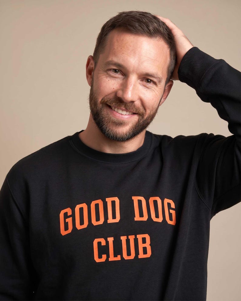 May include: A black long-sleeve sweatshirt featuring the text GOOD DOG CLUB printed in bold orange capital letters across the chest. The garment has a classic crew neckline and long sleeves, constructed from a solid black fabric material.