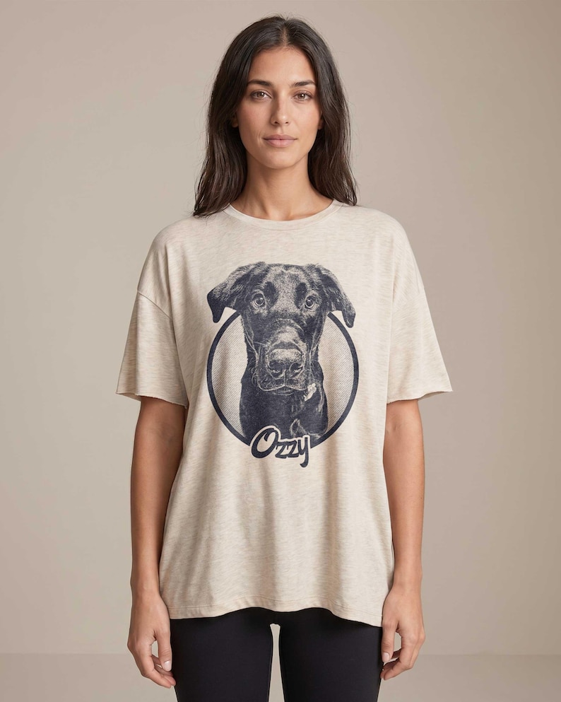 Puede incluir: Camiseta beige con una ilustraci&oacute;n en blanco y negro de un perro dentro de un c&iacute;rculo. El nombre del perro, "Ozzy", est&aacute; escrito en cursiva debajo de la imagen. La camiseta tiene mangas cortas y un corte holgado.