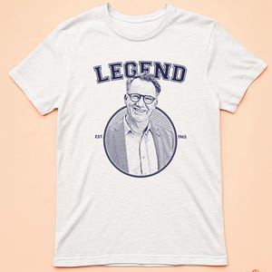 May include: White t-shirt with a blue graphic of a man wearing glasses and the text "LEGEND EST. 1965".