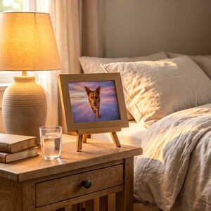 A small framed dog portrait propped on a wooden nightstand beside a lit lamp, with a made bed and layered cream pillows behind it.