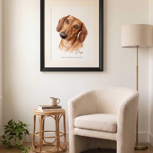 May include: A framed watercolor portrait of a brown dachshund dog with the name Ralph and the text Part of our family since 2022 printed below the illustration. The artwork is displayed in a black frame with a white mat on a light wall.