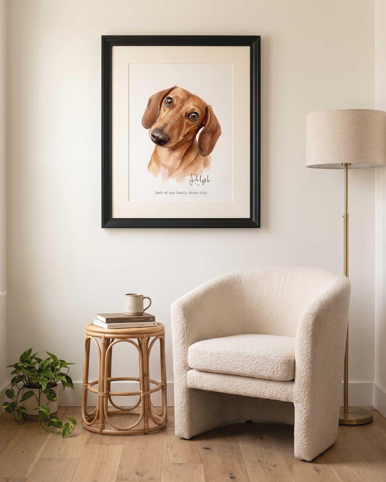 May include: A framed watercolor portrait of a brown dachshund dog with the name Ralph and the text Part of our family since 2022 printed below the illustration. The artwork is displayed in a black frame with a white mat on a light wall.