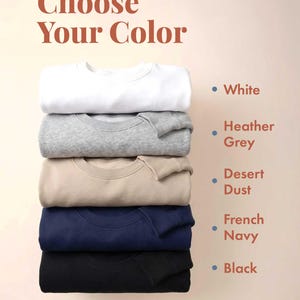 May include: A stack of five crewneck sweatshirts in white, heather grey, desert dust, french navy, and black. The text "Choose Your Color" appears at the top, with color names listed next to corresponding dots matching each sweatshirt color.