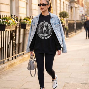 May include: A black sweatshirt featuring a circular graphic of a spaniel dog with the name CHARLIE printed below it. The sweatshirt is worn with a light blue denim jacket, black leggings, white sneakers, and a gray handbag.