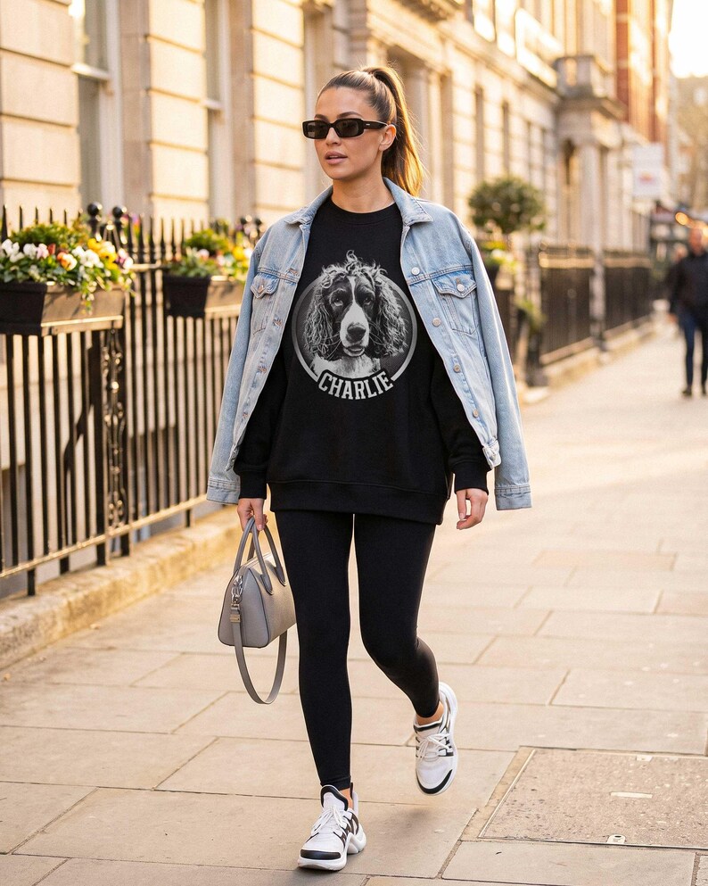 May include: A black sweatshirt featuring a circular graphic of a spaniel dog with the name CHARLIE printed below it. The sweatshirt is worn with a light blue denim jacket, black leggings, white sneakers, and a gray handbag.
