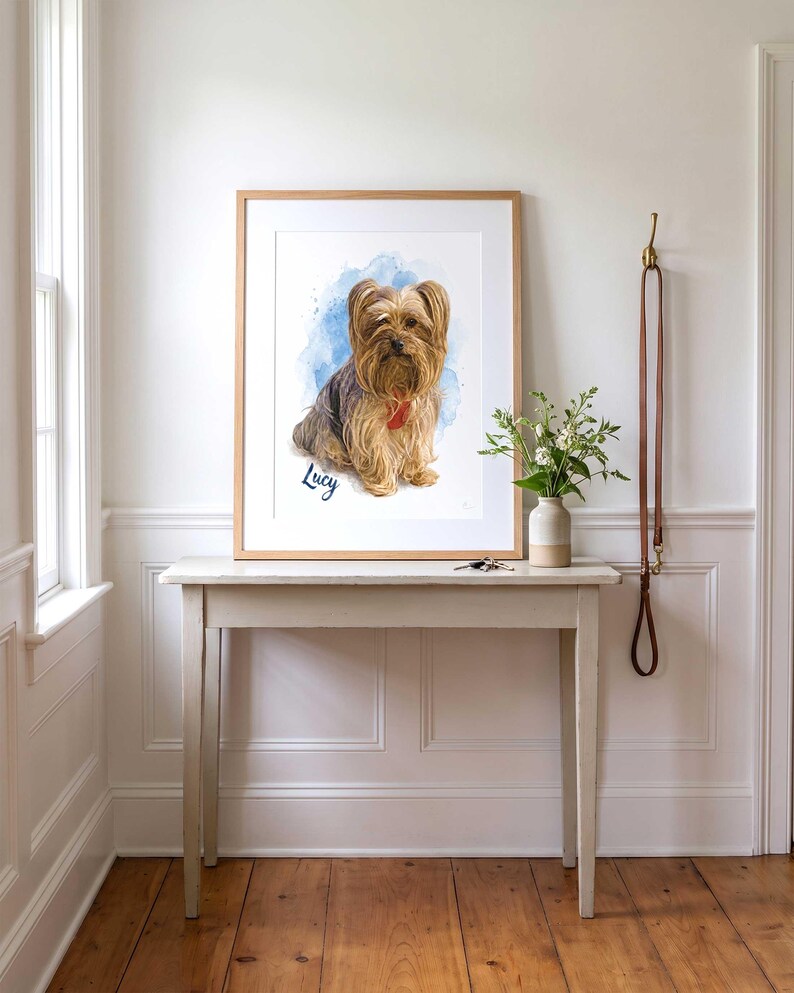 May include: A framed portrait print featuring a watercolor illustration of a Yorkshire Terrier dog with long tan and brown fur. The dog is set against a blue watercolor splash background with the name Lucy written in cursive script below the dog.