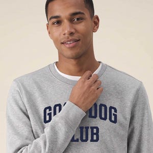 May include: A heather gray crewneck sweatshirt featuring the dark blue text GOOD DOG CLUB printed across the chest. The garment has a ribbed neckline and cuffs, with a white undershirt visible at the collar, providing a layered appearance.
