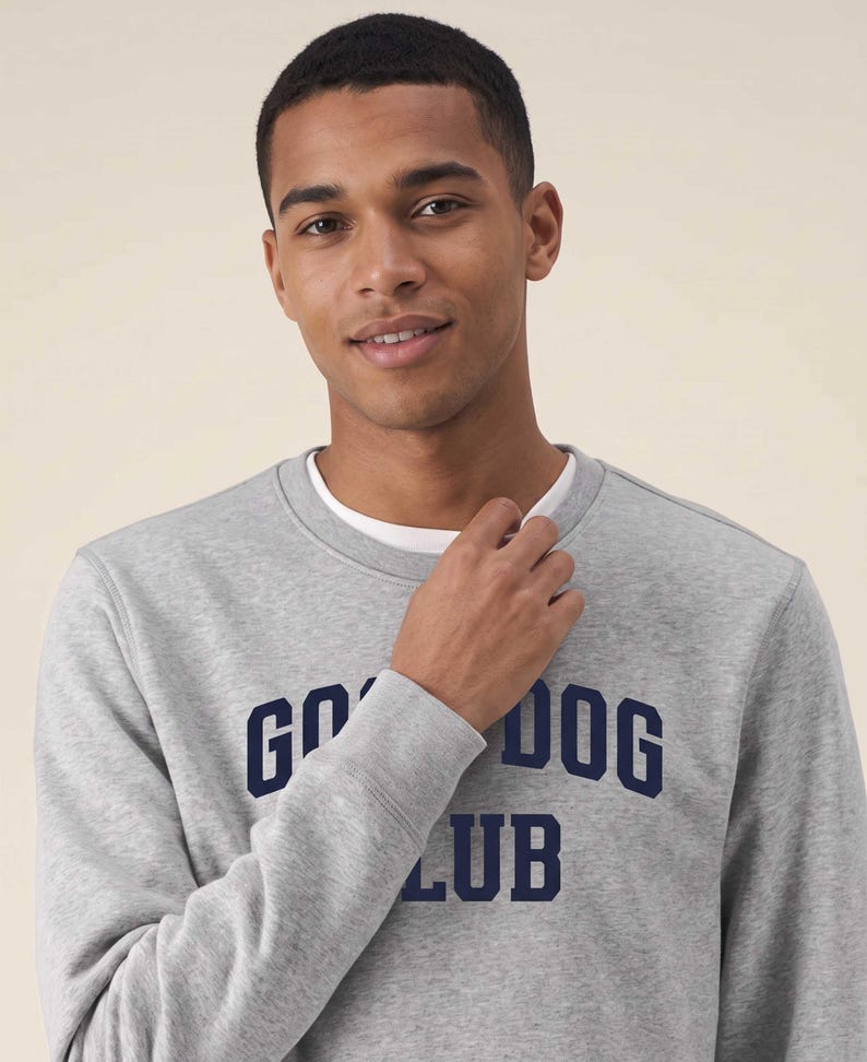May include: A heather gray crewneck sweatshirt featuring the dark blue text GOOD DOG CLUB printed across the chest. The garment has a ribbed neckline and cuffs, with a white undershirt visible at the collar, providing a layered appearance.