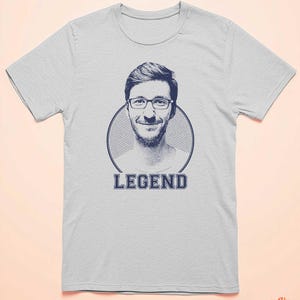 May include: A light grey t-shirt with a blue and white graphic of a man's face in a circle with the word "LEGEND" printed below the circle.