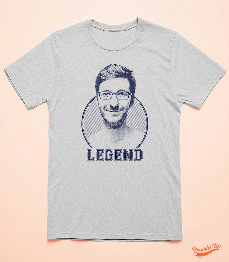 May include: A light grey t-shirt with a blue and white graphic of a man's face in a circle with the word "LEGEND" printed below the circle.