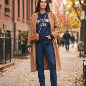 May include: A camel-colored wool coat worn over a navy blue sweatshirt with the text GOOD DOG CLUB in yellow lettering. The outfit includes dark blue denim jeans, a brown leather crossbody bag, and brown leather ankle boots on a city sidewalk.