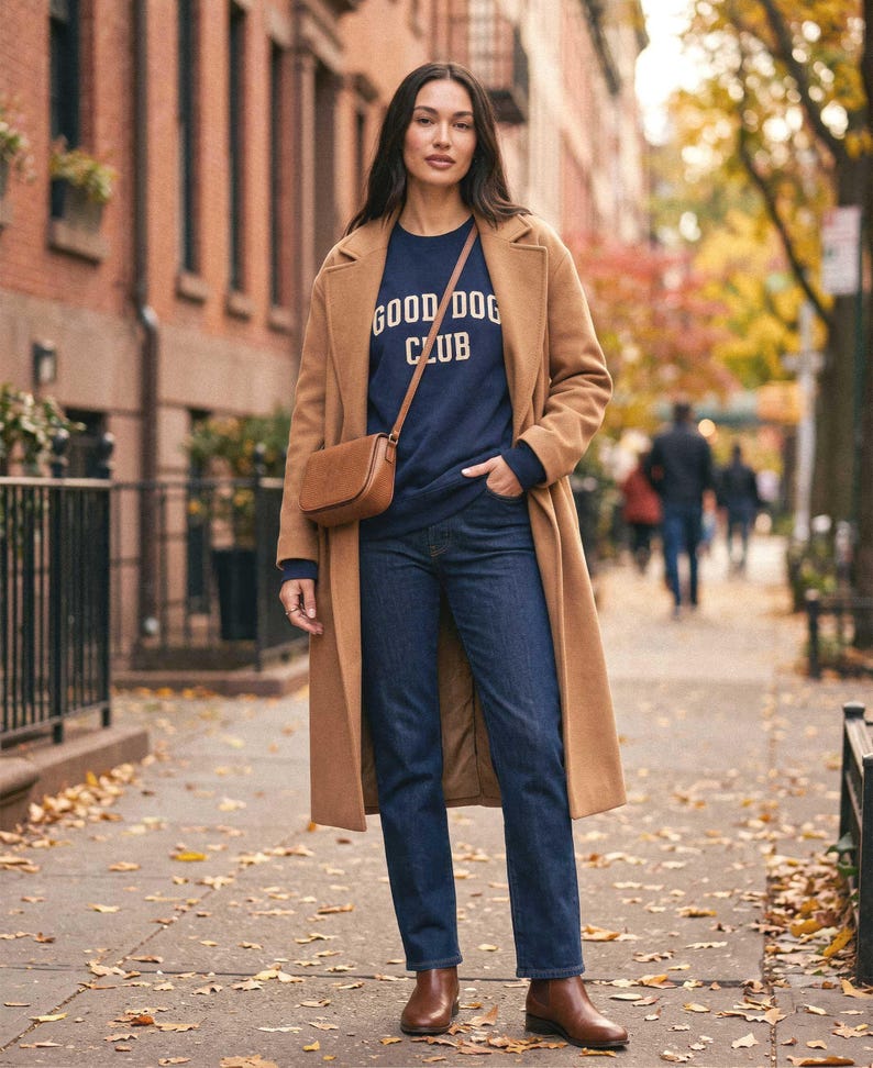 May include: A camel-colored wool coat worn over a navy blue sweatshirt with the text GOOD DOG CLUB in yellow lettering. The outfit includes dark blue denim jeans, a brown leather crossbody bag, and brown leather ankle boots on a city sidewalk.