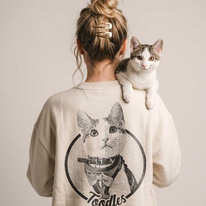 May include: A cream-colored long-sleeve sweatshirt featuring a black and white graphic print on the back. The print shows a cat wearing a harness and bow tie inside a circular frame with the text Toodles written in a script font below the cat.