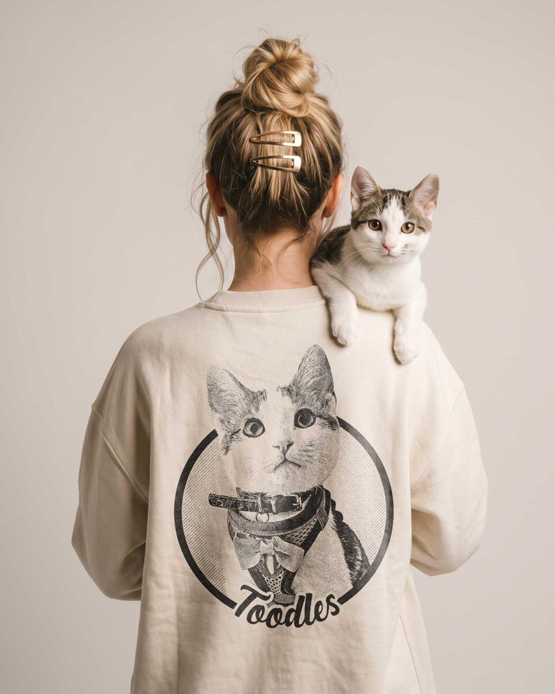May include: A cream-colored long-sleeve sweatshirt featuring a black and white graphic print on the back. The print shows a cat wearing a harness and bow tie inside a circular frame with the text Toodles written in a script font below the cat.