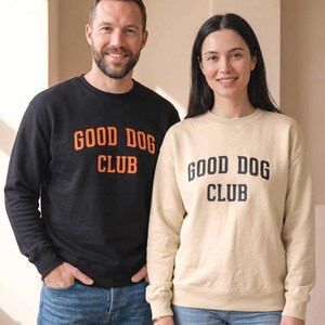 May include: A black crewneck sweatshirt with orange text reading GOOD DOG CLUB and a beige crewneck sweatshirt with black text reading GOOD DOG CLUB, both paired with blue denim jeans. The garments feature long sleeves and ribbed cuffs.