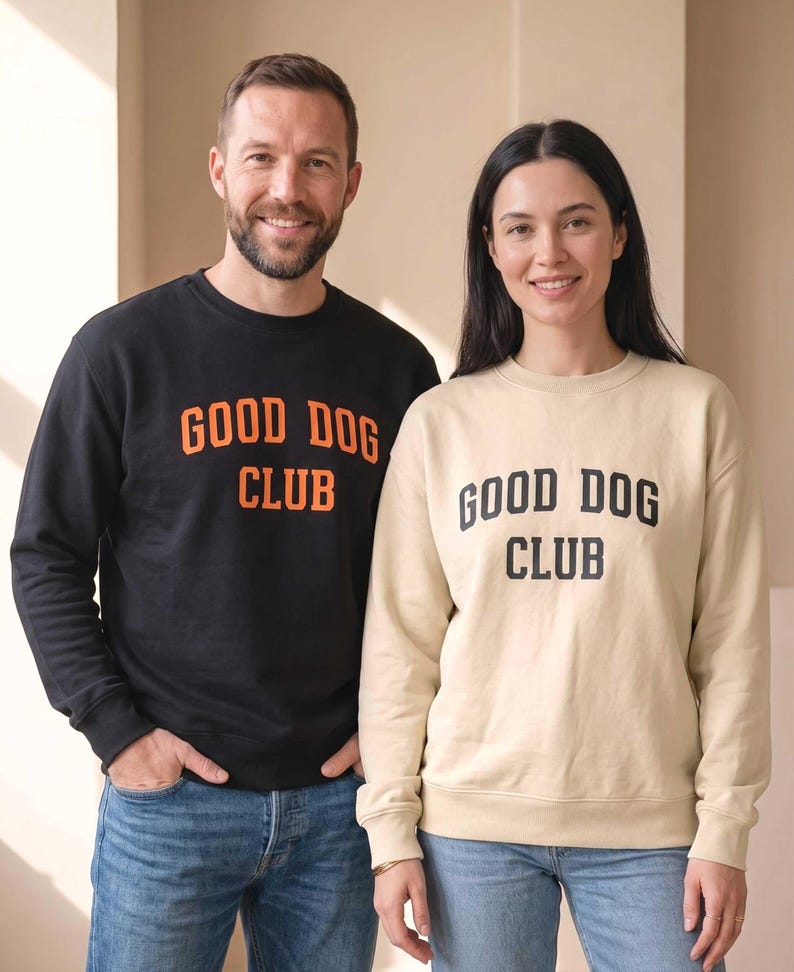 May include: A black crewneck sweatshirt with orange text reading GOOD DOG CLUB and a beige crewneck sweatshirt with black text reading GOOD DOG CLUB, both paired with blue denim jeans. The garments feature long sleeves and ribbed cuffs.