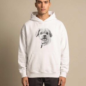 Puede incluir: Una sudadera con capucha blanca que presenta un boceto detallado en blanco y negro de la cara de un perro en el pecho. El perro tiene orejas ca&iacute;das y lleva un collar con una peque&ntilde;a placa. La sudadera incluye un bolsillo frontal y mangas largas.