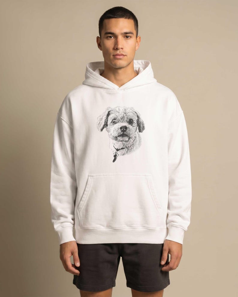 Puede incluir: Una sudadera con capucha blanca que presenta un boceto detallado en blanco y negro de la cara de un perro en el pecho. El perro tiene orejas ca&iacute;das y lleva un collar con una peque&ntilde;a placa. La sudadera incluye un bolsillo frontal y mangas largas.