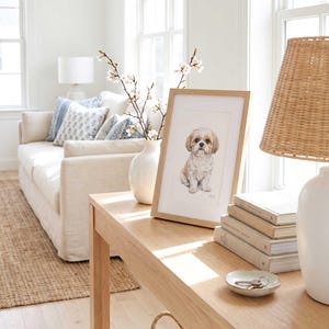 Two framed pastel pet portraits propped on a pale wood console against a white wall, beside a table lamp, a woven basket and a vase of dried stems, with a light sofa in the foreground.