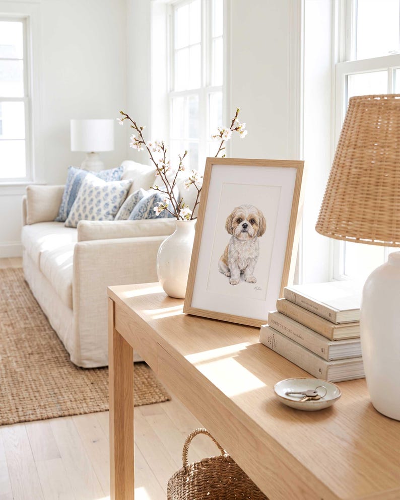 Two framed pastel pet portraits propped on a pale wood console against a white wall, beside a table lamp, a woven basket and a vase of dried stems, with a light sofa in the foreground.