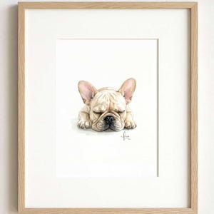 A framed watercolor illustration of a sleeping cream-colored French Bulldog with large ears and a black nose. The artwork is centered within a light-toned wooden frame, featuring a signature in the bottom right corner of the painting.