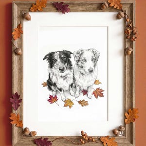 A framed charcoal-style drawing of two dogs sitting together, surrounded by colorful autumn leaves. The wooden frame is decorated with small acorns and oak leaves in shades of orange, red, and brown, creating a seasonal display.