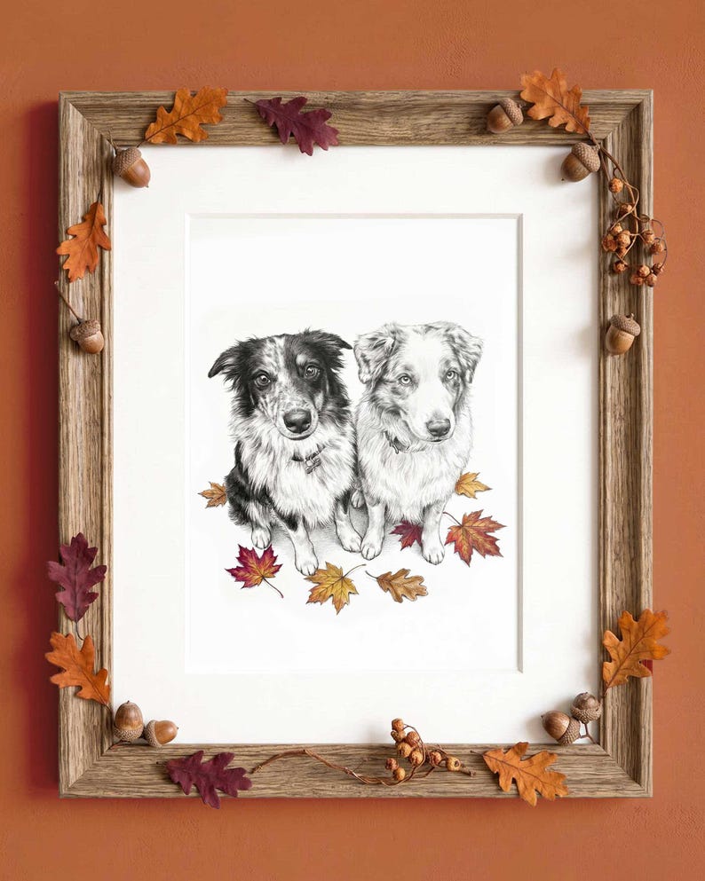 A framed charcoal-style drawing of two dogs sitting together, surrounded by colorful autumn leaves. The wooden frame is decorated with small acorns and oak leaves in shades of orange, red, and brown, creating a seasonal display.