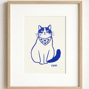 May include: A framed art print featuring a blue ink illustration of a cat wearing a patterned bandana with hearts and flowers. The cat is sitting upright and has the name Pippi written in blue text below its tail on a cream-colored background.