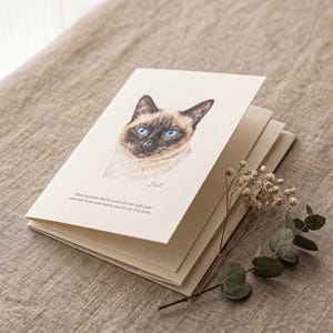 A pastel cat portrait printed on a folded cream card, lying on soft natural linen with a sprig of eucalyptus and a second blank card layered beneath it.