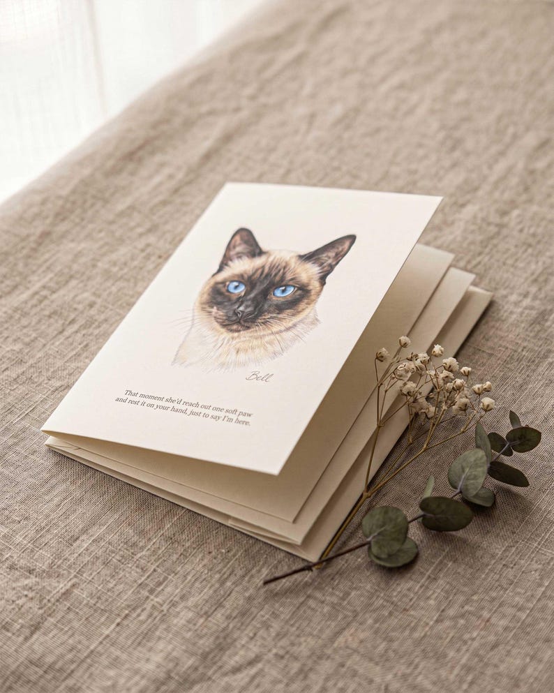 A pastel cat portrait printed on a folded cream card, lying on soft natural linen with a sprig of eucalyptus and a second blank card layered beneath it.