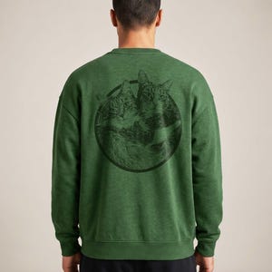May include: A forest green long-sleeve crewneck sweatshirt featuring a circular black graphic on the back. The graphic depicts two detailed cat faces side-by-side within the circle. The sweatshirt has a ribbed collar, cuffs, and hem.