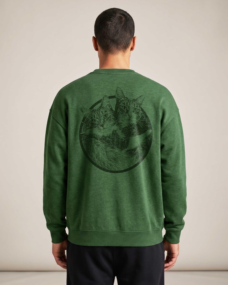 May include: A forest green long-sleeve crewneck sweatshirt featuring a circular black graphic on the back. The graphic depicts two detailed cat faces side-by-side within the circle. The sweatshirt has a ribbed collar, cuffs, and hem.