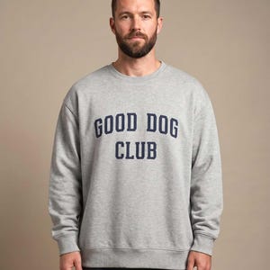 May include: A light gray crewneck sweatshirt featuring the text GOOD DOG CLUB printed in dark navy blue block lettering across the chest. The garment has long sleeves with ribbed cuffs and a ribbed hem, providing a standard fit.