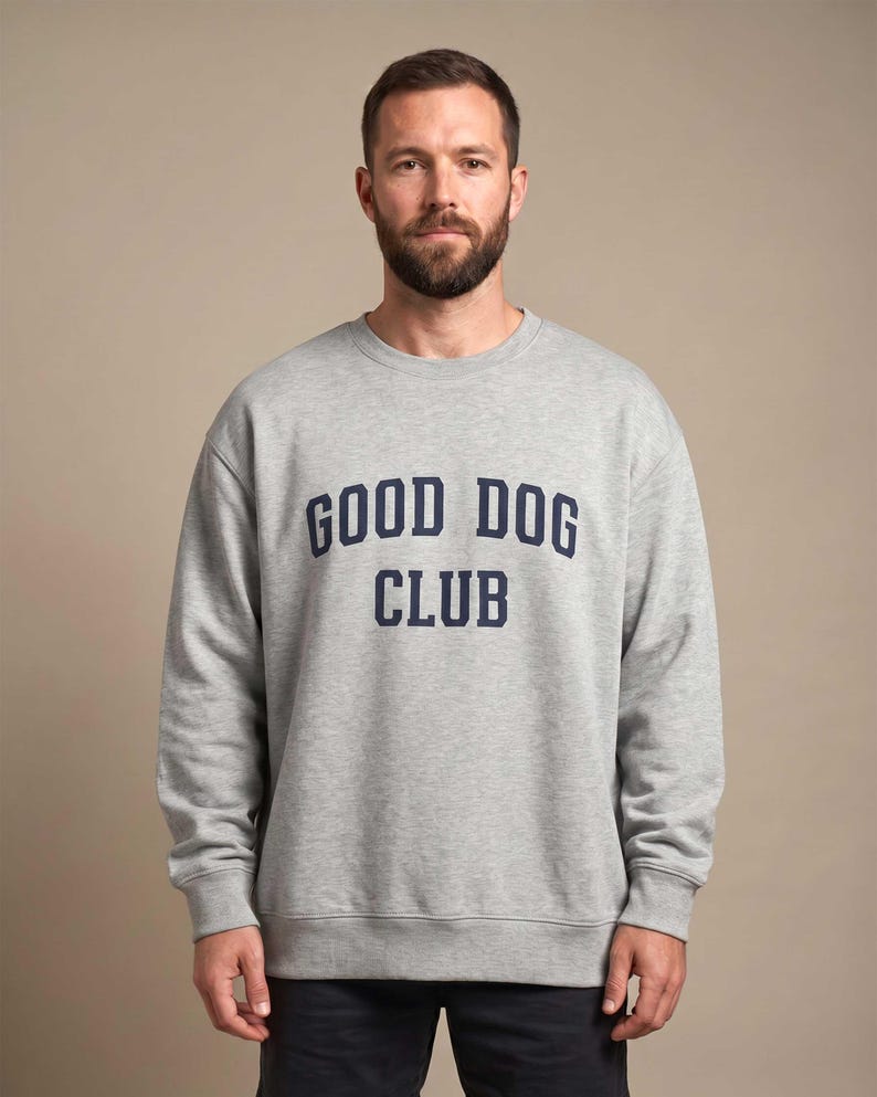 May include: A light gray crewneck sweatshirt featuring the text GOOD DOG CLUB printed in dark navy blue block lettering across the chest. The garment has long sleeves with ribbed cuffs and a ribbed hem, providing a standard fit.