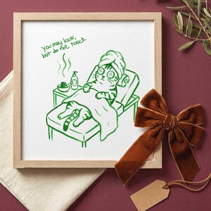 Op de afbeelding: Een vierkante ingelijste print met een groene lijntekening van een kat met een handdoek op het hoofd en komkommerschijfjes op de ogen, liggend op een spabank. De tekst luidt, You may look, but do not touch. De print zit in een licht houten lijst.