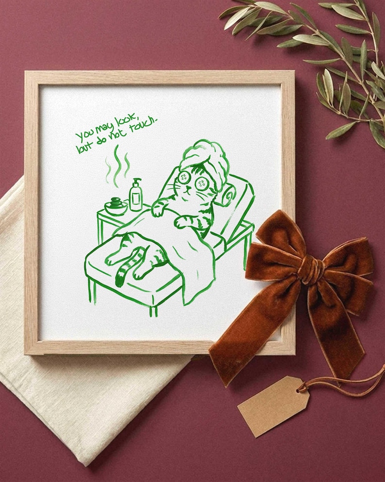 Op de afbeelding: Een vierkante ingelijste print met een groene lijntekening van een kat met een handdoek op het hoofd en komkommerschijfjes op de ogen, liggend op een spabank. De tekst luidt, You may look, but do not touch. De print zit in een licht houten lijst.