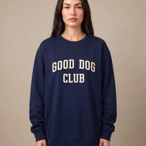 May include: A navy blue crewneck sweatshirt featuring the text GOOD DOG CLUB printed in a cream-colored, collegiate-style font across the chest. The garment has long sleeves with ribbed cuffs and a ribbed hem, providing a standard fit.