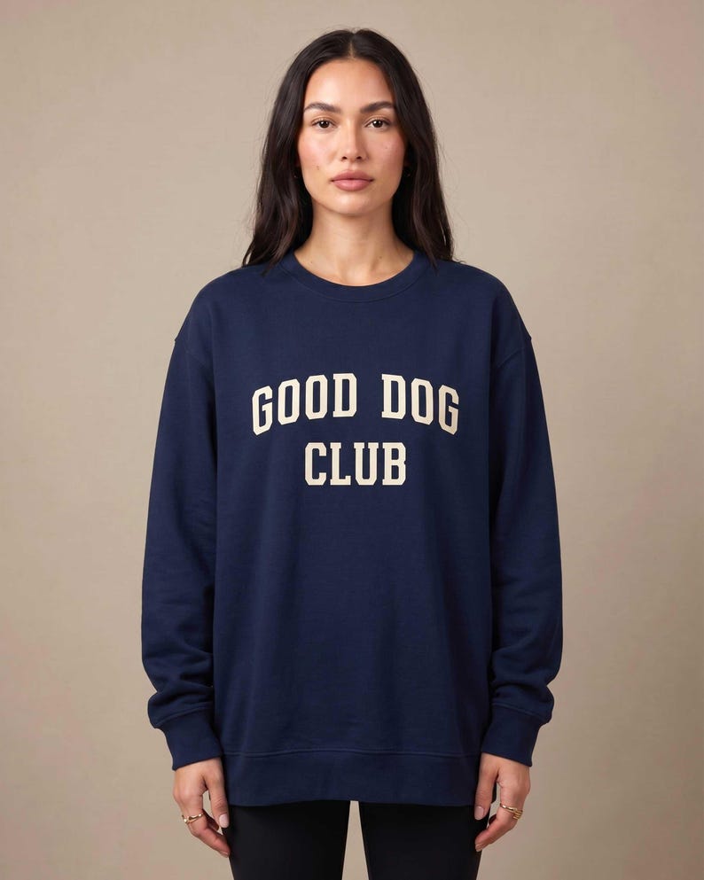 May include: A navy blue crewneck sweatshirt featuring the text GOOD DOG CLUB printed in a cream-colored, collegiate-style font across the chest. The garment has long sleeves with ribbed cuffs and a ribbed hem, providing a standard fit.