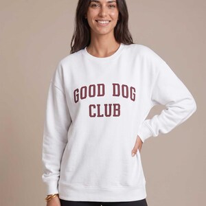 May include: A white long-sleeve crewneck sweatshirt featuring the text GOOD DOG CLUB printed in a dark maroon collegiate-style font across the chest. The garment has ribbed cuffs and a ribbed hem, providing a structured fit for the torso and arms.