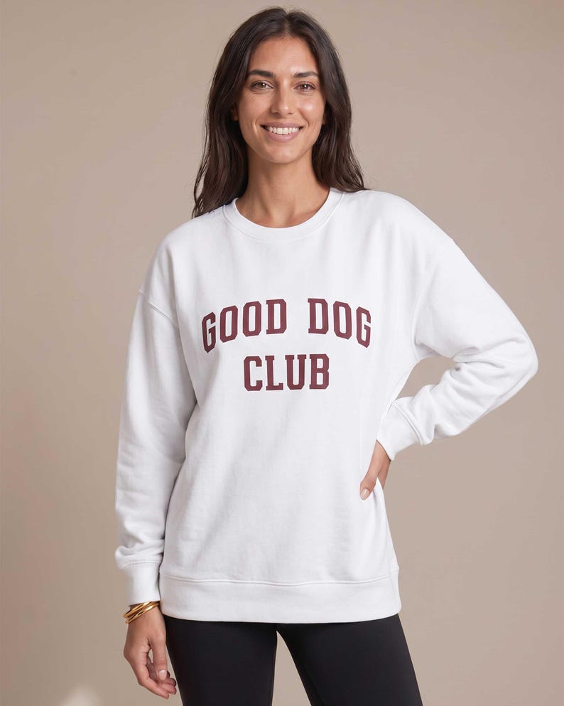 May include: A white long-sleeve crewneck sweatshirt featuring the text GOOD DOG CLUB printed in a dark maroon collegiate-style font across the chest. The garment has ribbed cuffs and a ribbed hem, providing a structured fit for the torso and arms.