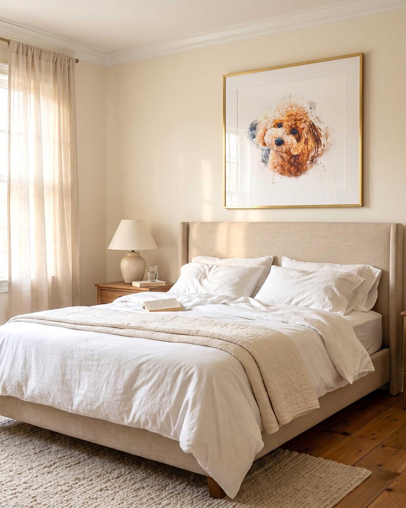 May include: A square framed wall art piece featuring a watercolor-style portrait of a brown curly-haired dog. The artwork is displayed in a thin gold frame with a white mat, hanging on a light beige wall above a neutral-toned upholstered bed.