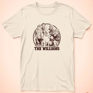 May include: A beige t-shirt featuring a brown circular graphic of a family and their dog. The text "THE WILLIAMS" is printed beneath the circle.