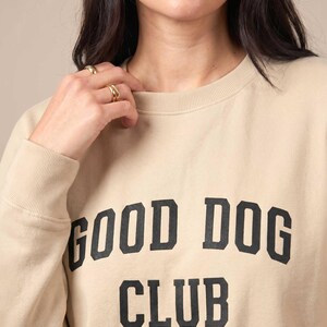 May include: A beige crewneck sweatshirt featuring the text GOOD DOG CLUB printed in a bold, black, collegiate-style font across the chest. The garment has long sleeves with ribbed cuffs and a ribbed neckline, providing a simple and clean design.