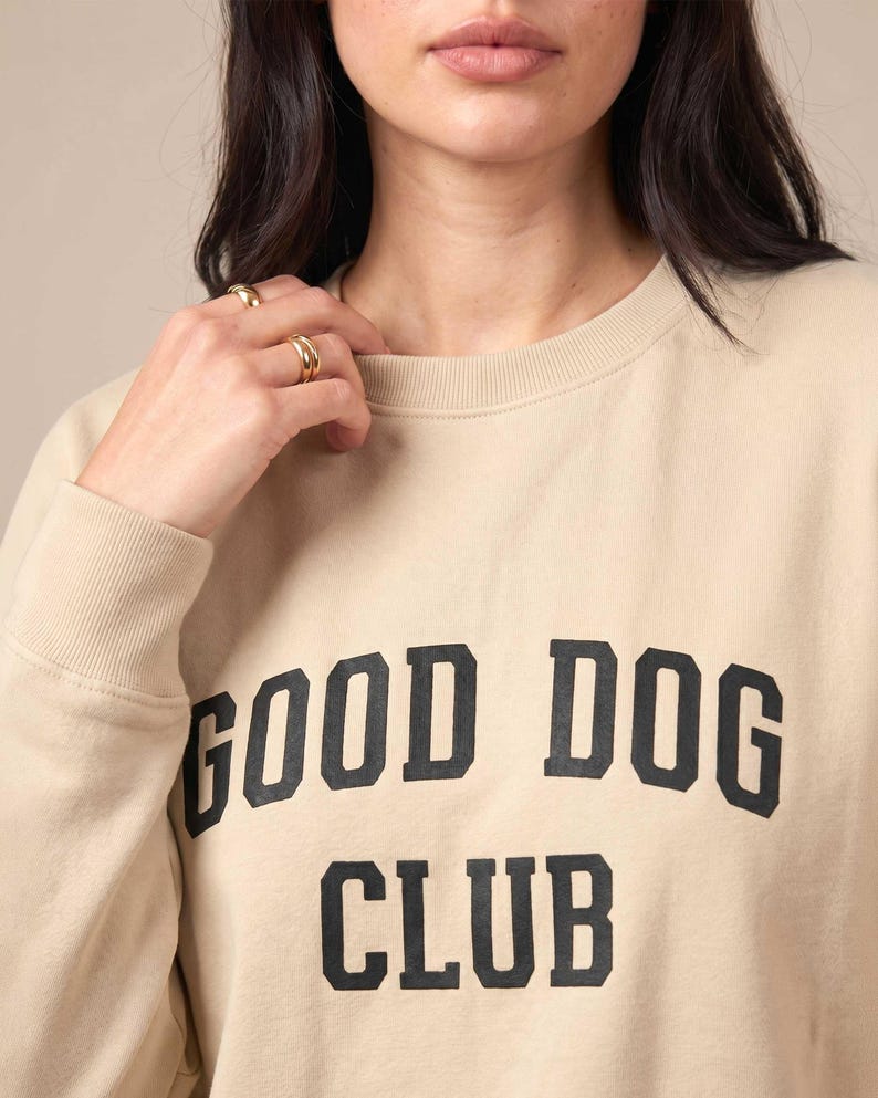 May include: A beige crewneck sweatshirt featuring the text GOOD DOG CLUB printed in a bold, black, collegiate-style font across the chest. The garment has long sleeves with ribbed cuffs and a ribbed neckline, providing a simple and clean design.