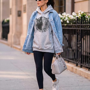 Puede incluir: Una sudadera con capucha gris claro que presenta un gr&aacute;fico circular de un gato negro y un gato atigrado con el texto Panther & Kitty, usada bajo una chaqueta de mezclilla azul claro con leggings negros, zapatillas blancas y un bolso gris.