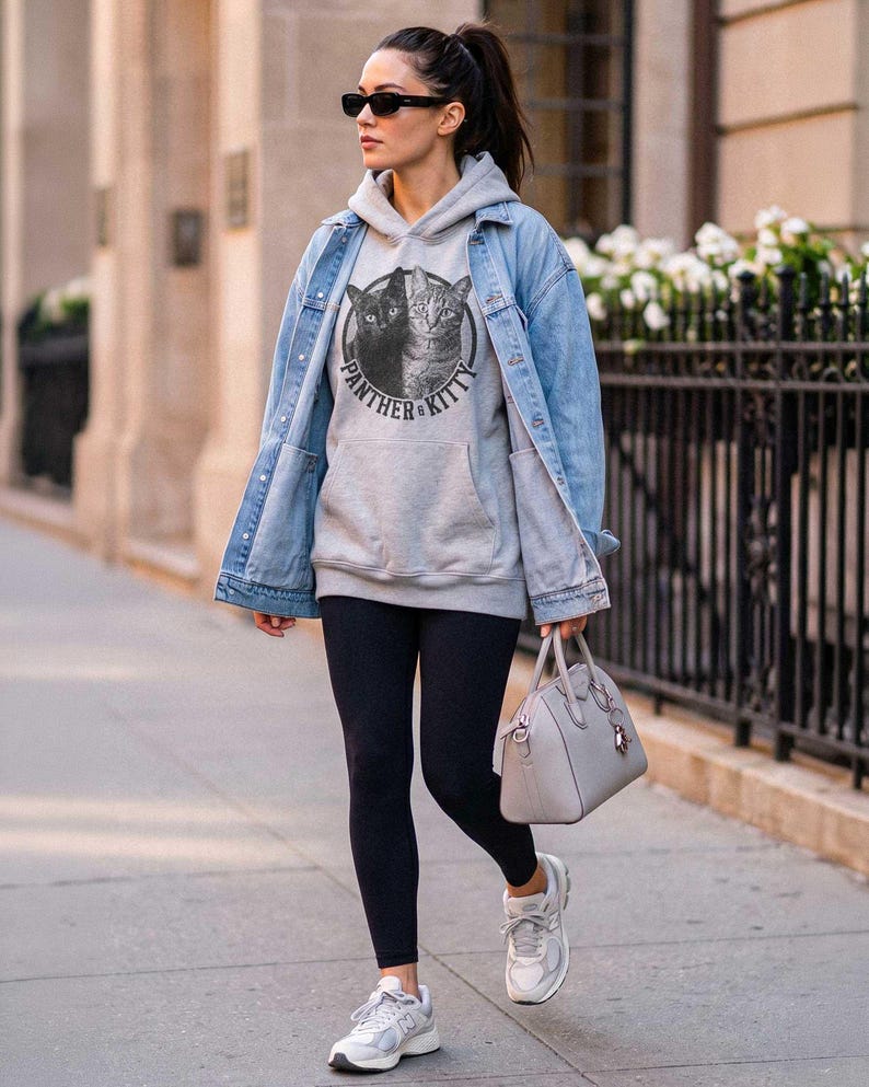 Puede incluir: Una sudadera con capucha gris claro que presenta un gr&aacute;fico circular de un gato negro y un gato atigrado con el texto Panther & Kitty, usada bajo una chaqueta de mezclilla azul claro con leggings negros, zapatillas blancas y un bolso gris.